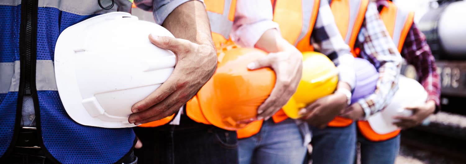 Workers stand in a line wearing safety vests and holding hard hats