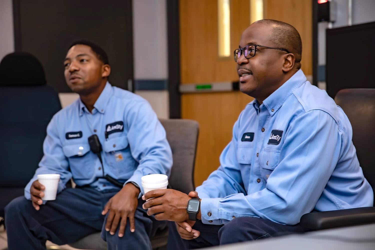 Two employees hold foam coffee cups while seated in a break room