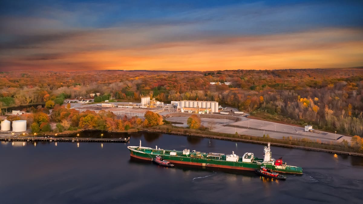 A barge prepares to dock at a river port surrounded by trees with fall foliage