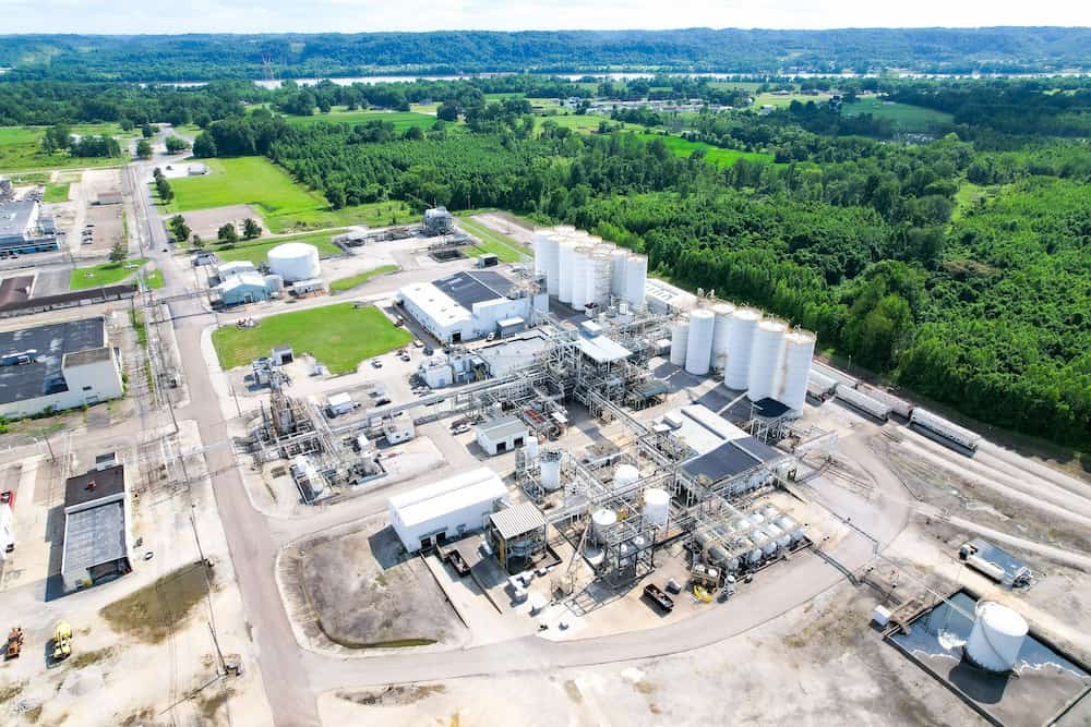 An aerial view of a large manufacturing facility surrounded by green trees with a river behind it.